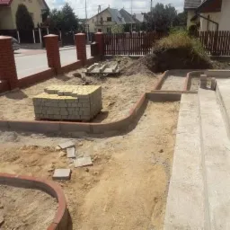 Przygotowanie terenu pod układanie kostki brukowej: paleta żółtej kostki, obrzeża z czerwonej kostki klinkierowej, widoczne fundamenty schodów i częściowo wykopany teren.