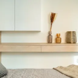 Minimalistyczna sypialnia z drewnianą konsolą i dekoracjami: szklane wazony z suszonymi trawami, metalowa lampa, białe szafki wiszące i szara pościel z poduszką.