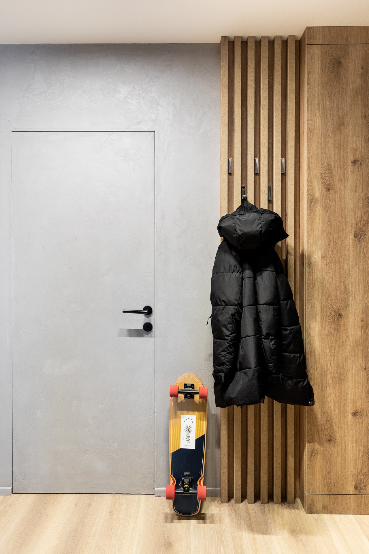 Nowoczesny przedpokój z szarą, gładką ścianą, ukrytymi drzwiami, drewnianym panelem z wieszakami, na którym wisi czarna kurtka puchowa i deskorolką typu cruiser z motywem palmy opartą o ścianę.