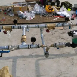 Elementy instalacji z rur, zaworów, manometrów i pompy obiegowej zamontowane na metalowym stelażu, widoczne narzędzia i akcesoria instalacyjne w tle.