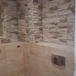 Wykończona ściana łazienki z imitacją kamienia na górze i beżowymi płytkami ceramicznymi na dole, widoczny fragment lustra po lewej stronie i metalowy panel sterujący spłuczką.