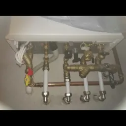 Podwieszony piec gazowy z widocznymi rurami miedzianymi i białymi plastikowymi rurami, oraz zaworami, zainstalowany w jasnej wnęce.