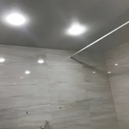 Wykończona łazienka z szarymi płytkami imitującymi marmur, oświetleniem punktowym w suficie i zamontowanym białym drążkiem na zasłonę prysznicową, widoczne ślady po wierceniu.