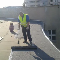 Pracownik w kamizelce odblaskowej zgrzewa papę na płaskim dachu budynku, widoczna butla gazowa i narzędzia dekarskie.