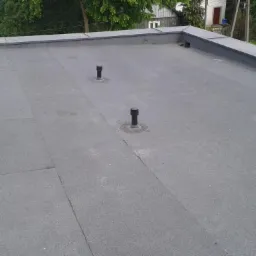 Płaski dach pokryty papą z trzema pionowymi kominkami wentylacyjnymi, widok z góry na tle zieleni i zabudowań.