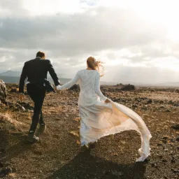 Para młoda, on w czarnym garniturze, ona w białej sukni ślubnej, idąca po skalistym terenie w kierunku zachodzącego słońca, z powiewającą suknią i wiatrem we włosach.