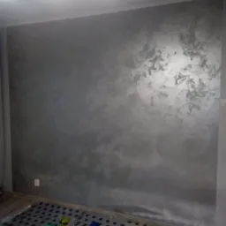 Ściana w pokoju pokryta dekoracyjną, srebrzysto-szarą farbą z efektem chmur, zabezpieczona folią ochronną na podłodze.