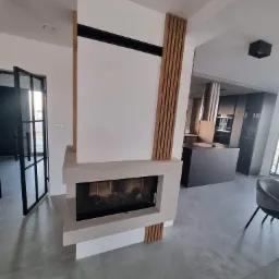 Nowoczesny salon z kominkiem obudowanym płytami imitującymi beton, drewniane panele dekoracyjne po bokach kominka, szklane drzwi w czarnej ramie oraz widoczna w tle wyspa kuchenna z okapem.