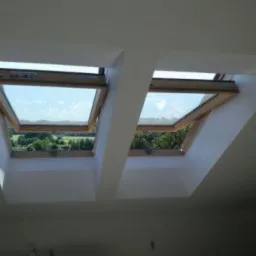 Dwa otwarte okna dachowe z drewnianymi ramami w białym, świeżo wykończonym poddaszu, widok na zieleń na zewnątrz.