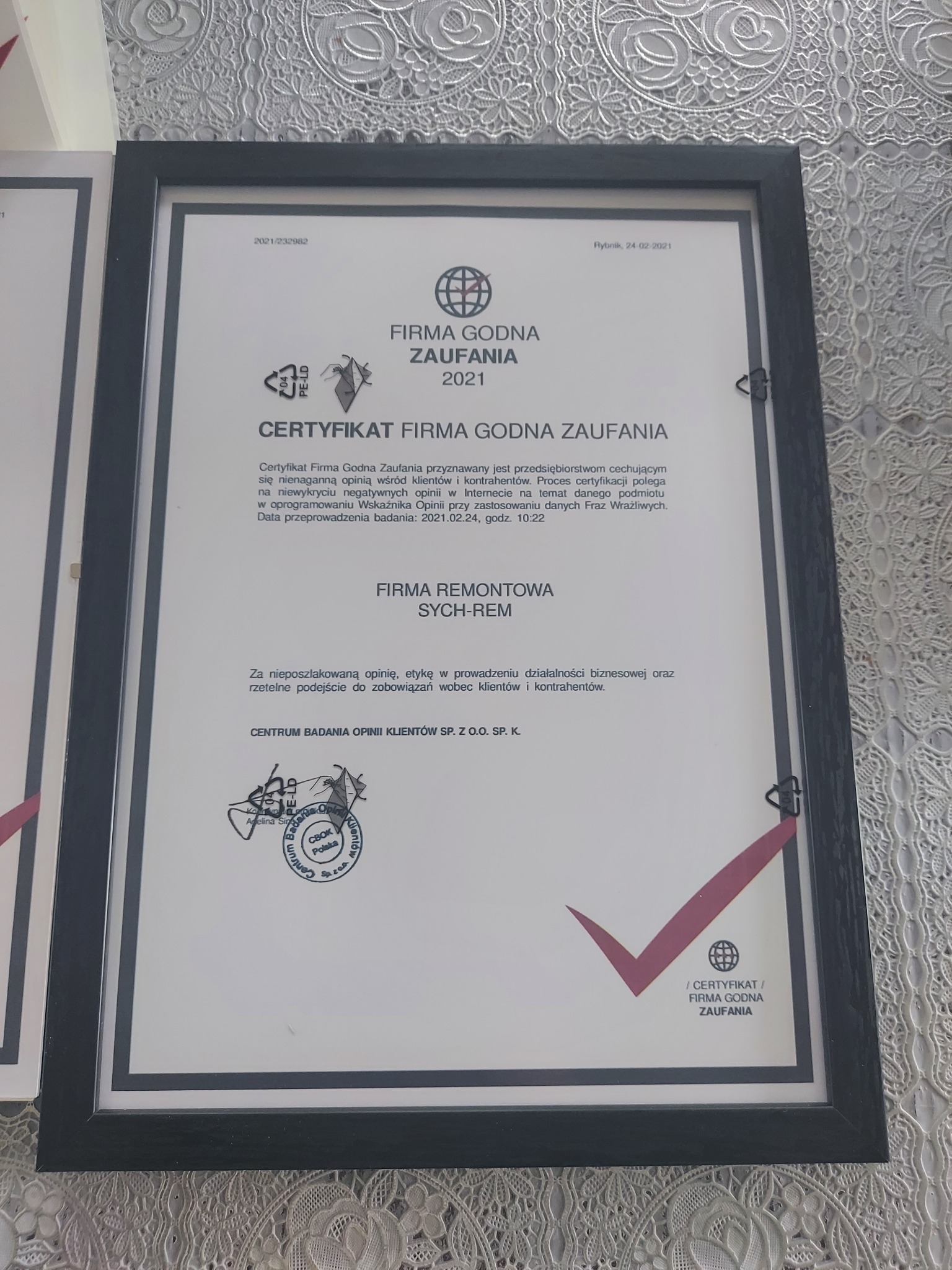 Oprawiony w ramkę certyfikat 'Firma Godna Zaufania 2021' firmy remontowej Sych-Rem, wiszący na ścianie z wzorzystą tapetą.