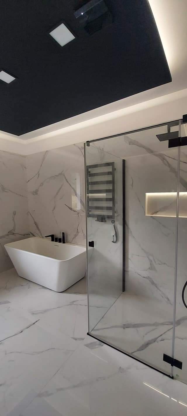 Nowoczesna łazienka z białą wanną wolnostojącą, kabiną prysznicową ze szklanymi drzwiami przesuwnymi i czarnym grzejnikiem drabinkowym na ścianie z marmurowym wzorem.