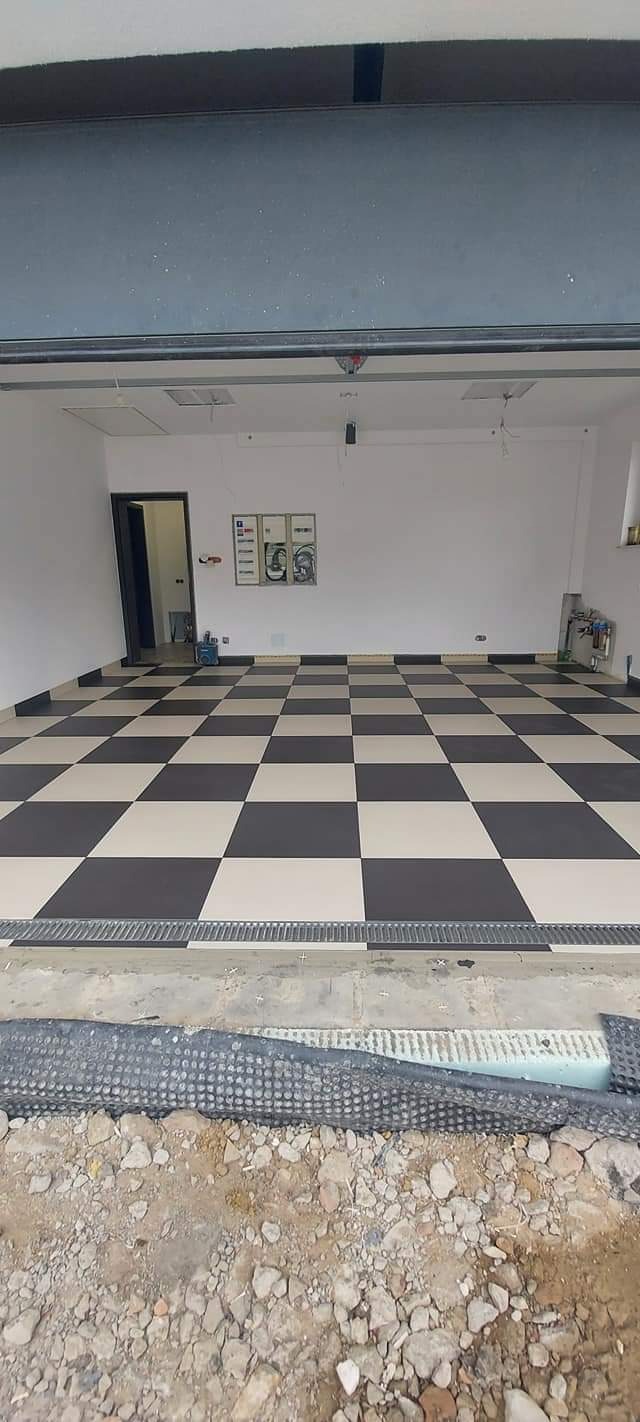 Wykończone wnętrze garażu z czarno-białą szachownicą na podłodze, widoczne instalacje elektryczne na ścianie, otwarte drzwi do pomieszczenia gospodarczego i fragment podłoża przed garażem...