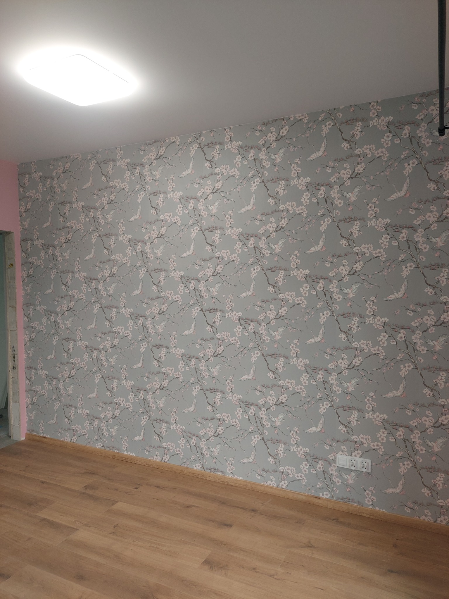Ściana z szarym, kwiecistym wzorem tapety z motywem żurawi i drewniana podłoga z panelami, widoczne gniazdka elektryczne.