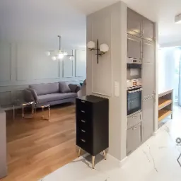 Nowoczesne wnętrze mieszkania z szarą sofą, drewnianą podłogą i zabudową kuchenną w odcieniach szarości, oświetlone lampami kulami i ozdobione sztukaterią.