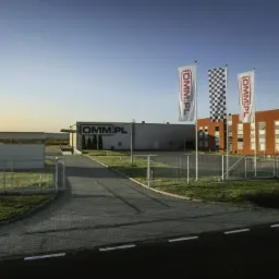 Nowoczesny budynek biurowo-produkcyjny z logo firmy na elewacji i flagach, otoczony zielenią i ogrodzeniem, widok z zewnątrz przy słonecznej pogodzie.