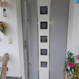 Szare drzwi zewnętrzne z długim, pionowym uchwytem i pięcioma kwadratowymi okienkami, widoczne z zewnątrz domu, z dekoracjami kwiatowymi po prawej stronie i fragmentem drewnianej ławki po lewej.