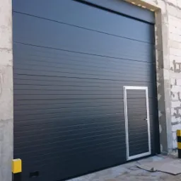 Zamknięta, grafitowa brama segmentowa z wbudowanymi drzwiami, widoczna na tle surowej ściany z białego pustaka i betonowych filarów, z żółto-czarnym słupkiem zabezpieczającym po lewej stronie.