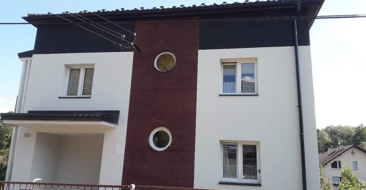 Dwupiętrowy dom z białą elewacją, ciemnobrązową wstawką z okrągłymi oknami i czarnym dachem, widoczny z zewnątrz.