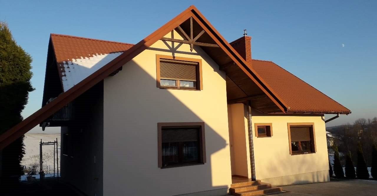 Dwukondygnacyjny dom z brązowym dachem, okna z roletami zewnętrznymi i elewacją w odcieniach beżu, oświetlony popołudniowym słońcem, częściowo pokryty śniegiem.