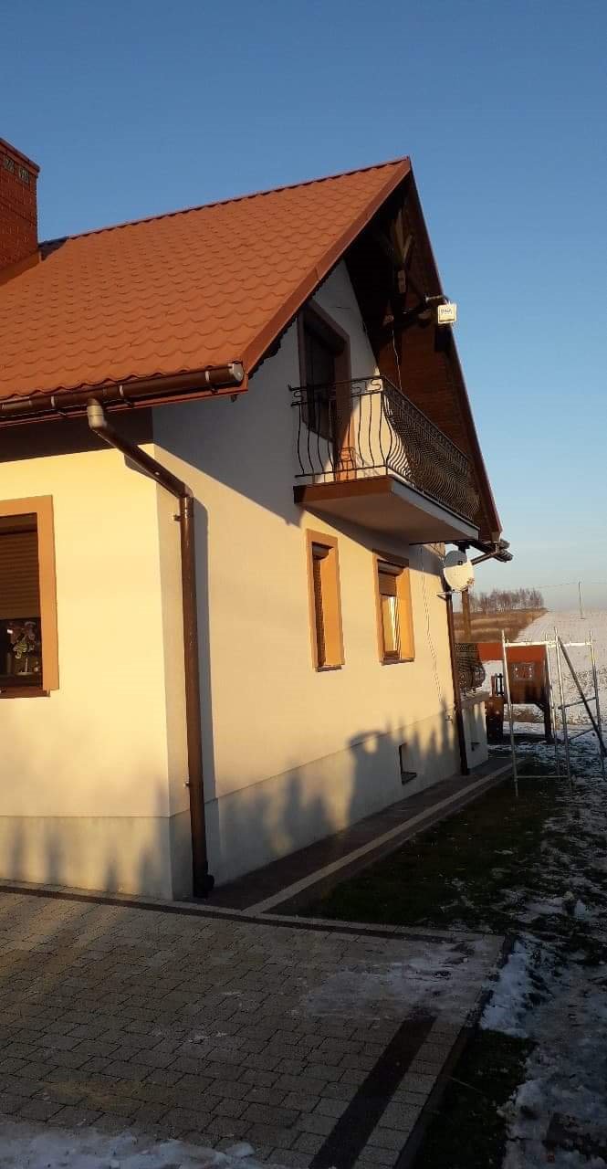 Dwupiętrowy dom z beżową elewacją, brązowymi oknami i balkonem z kutą balustradą, sfotografowany w zimowym krajobrazie, z widocznym fragmentem chodnika z kostki brukowej i resztkami śniegu.