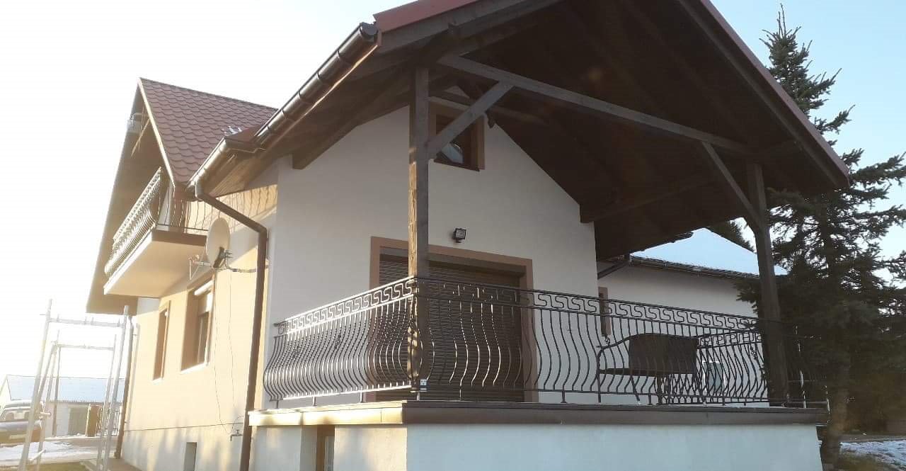 Jasna elewacja domu jednorodzinnego z brązowymi akcentami, widoczny balkon z ozdobną metalową balustradą i drewniana konstrukcja zadaszenia tarasu.
