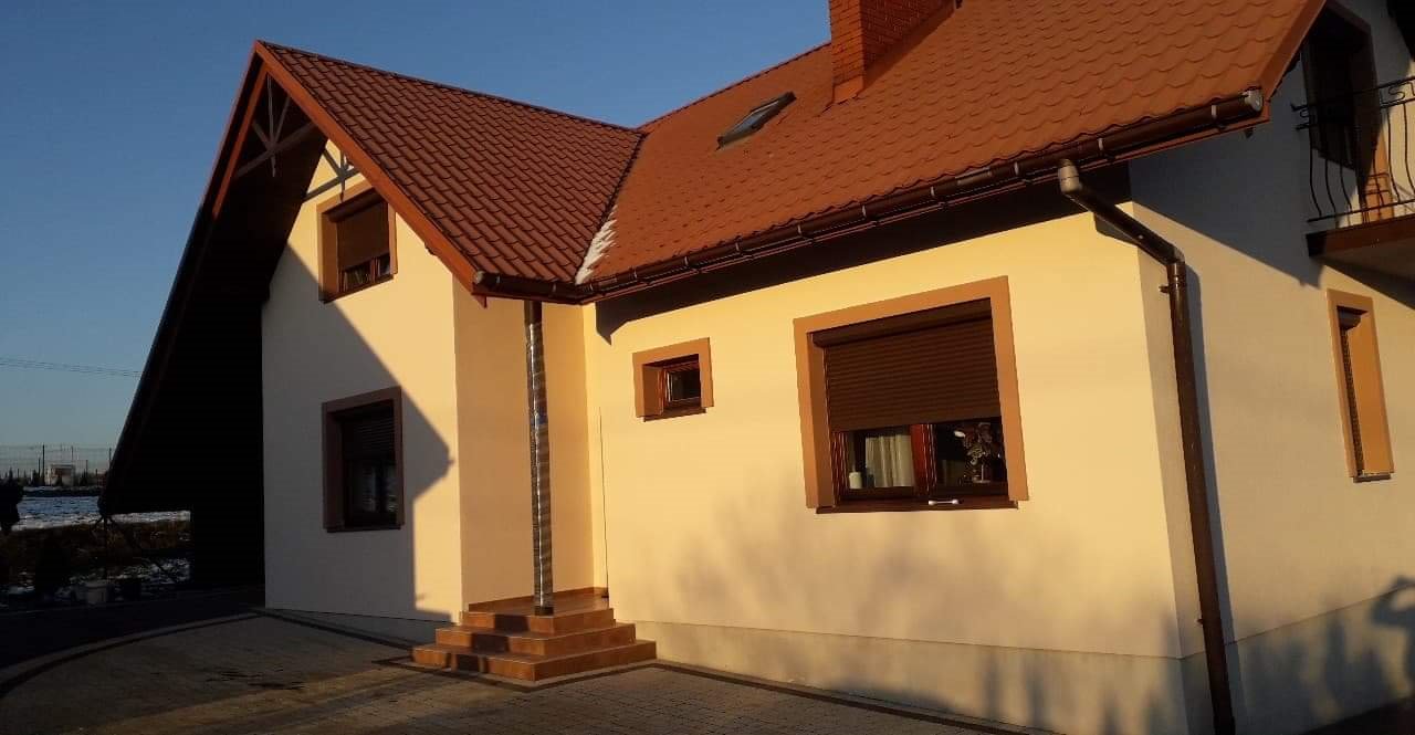 Dom jednorodzinny w Krzeszowicach z ocieploną i pomalowaną elewacją w odcieniach beżu, z brązowymi oknami z roletami zewnętrznymi i brązową dachówką.