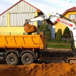 Żółta wywrotka i biała koparka Bobcat E80 w trakcie załadunku ziemi na tle dwóch żółtych domów jednorodzinnych.