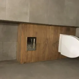 Nowoczesna toaleta podwieszana z drewnianą obudową i ukrytym systemem spłukiwania w łazience wykończonej szarymi płytkami.