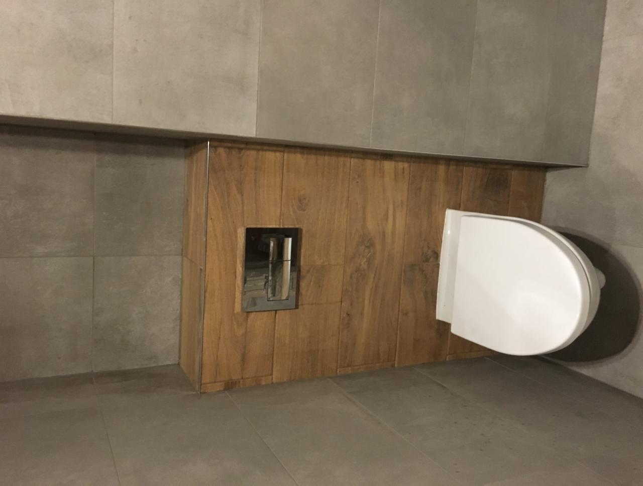 Nowoczesna toaleta podwieszana z drewnianą obudową i ukrytym systemem spłukiwania w łazience wykończonej szarymi płytkami.