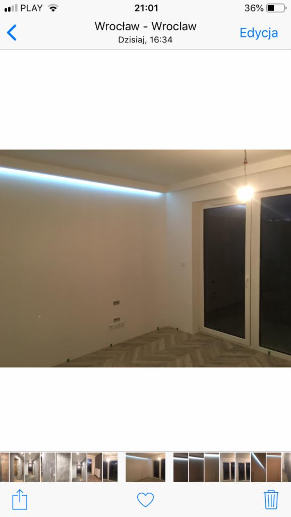 Wykończone wnętrze pokoju z jasnymi ścianami, podłogą ułożoną w jodełkę i oświetleniem LED przy suficie. W tle widoczne okna balkonowe.