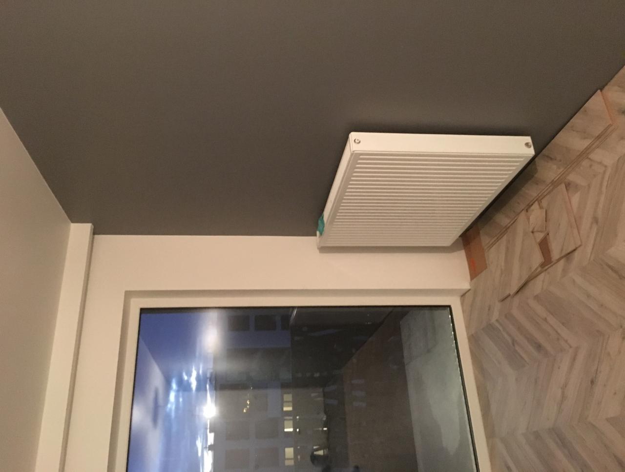 Szare wnętrze z zamontowanym białym grzejnikiem pod sufitem, widoczna część okna i podłoga z ułożonym parkietem w jodełkę.