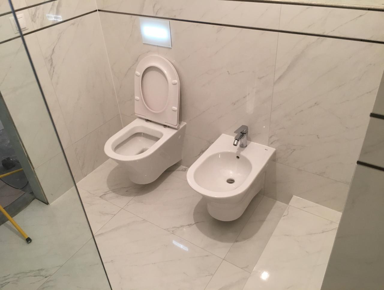 Nowoczesna łazienka z białymi marmurowymi płytkami, wiszącą toaletą z otwartą klapą i bidetem z chromowaną baterią, widoczne fragmenty lustra i drzwi.