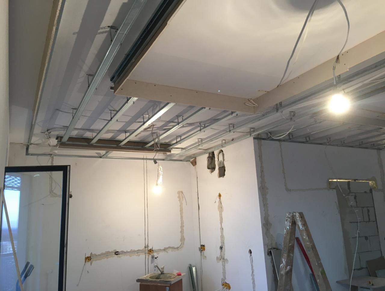 Montaż sufitu podwieszanego z widoczną metalową konstrukcją i częściowo zamontowanymi płytami gipsowymi, instalacja elektryczna w ścianach w trakcie prac.