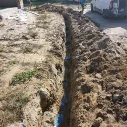 Wykop w ziemi z ułożonymi niebieskimi rurami, widoczni dwaj pracownicy w oddali i samochody dostawcze w tle.