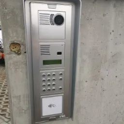 Srebrny panel domofonu Urmet z kamerą i czytnikiem kart zbliżeniowych zamontowany na surowym, betonowym słupku.