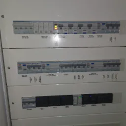 Rozdzielnica elektryczna z widocznymi bezpiecznikami, wyłącznikami różnicowoprądowymi, ogranicznikami przepięć i zasilaczami do fotokomórek, oznaczona etykietami w języku polskim.