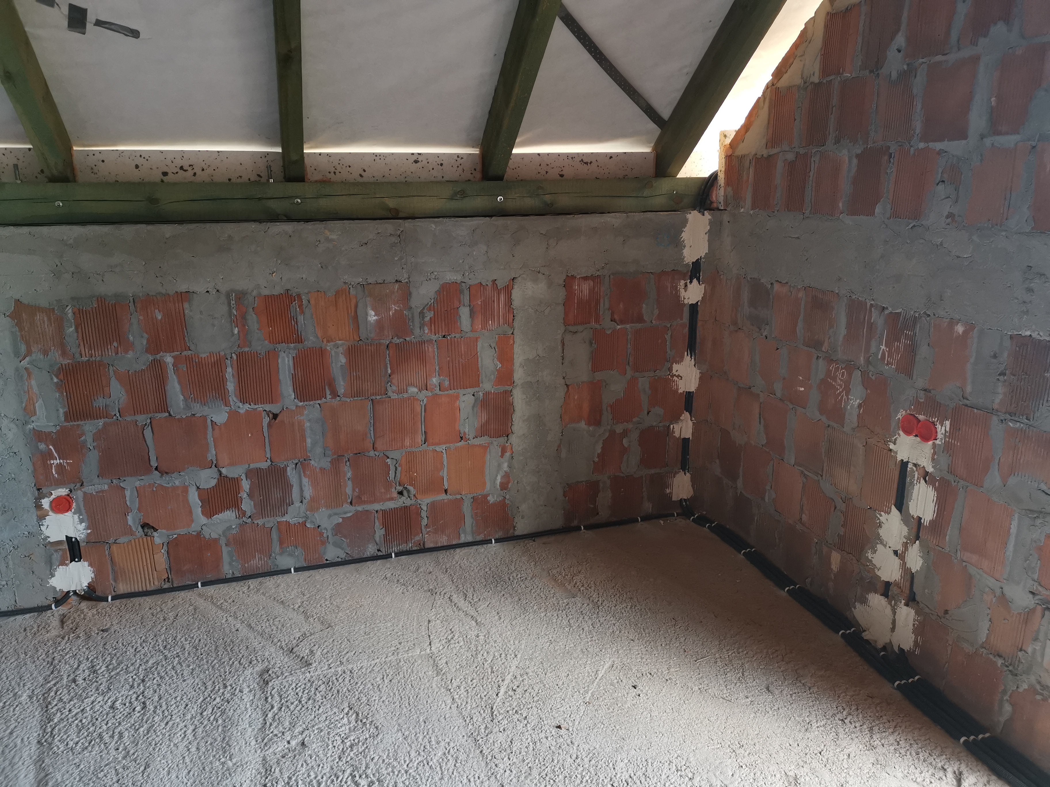 Wnętrze poddasza w trakcie instalacji elektrycznej: widoczne kable w peszlach mocowane do ceglanej ściany, puszki elektryczne pod tynk i drewniana konstrukcja dachu.