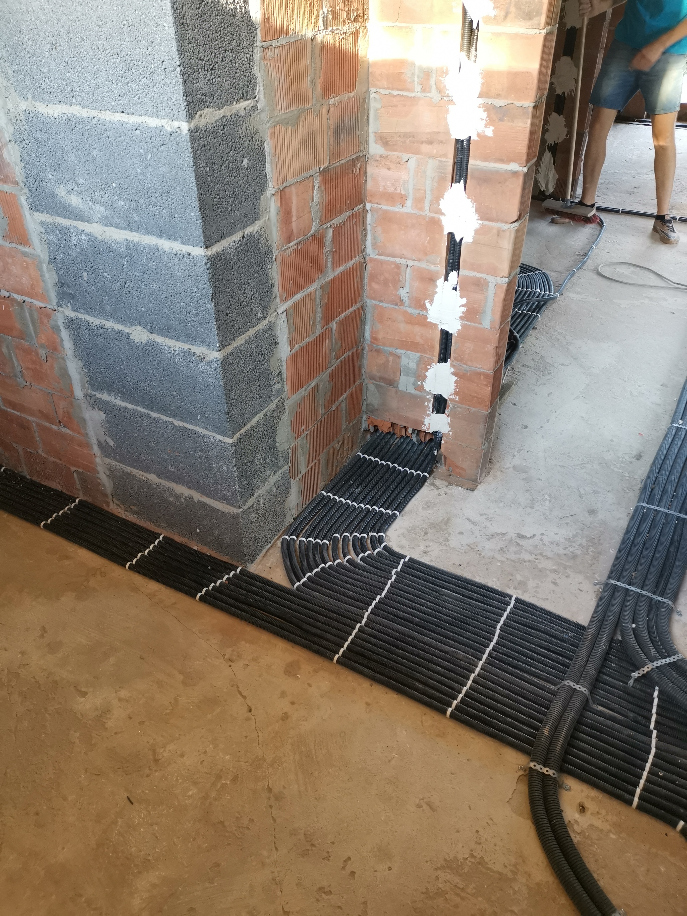 Rzędy czarnych, karbowanych rur ułożone równolegle na surowej podłodze, wchodzące w otwór w ścianie z czerwonej cegły i szarego betonu komórkowego, zabezpieczone białym gipsem w szczelinie.