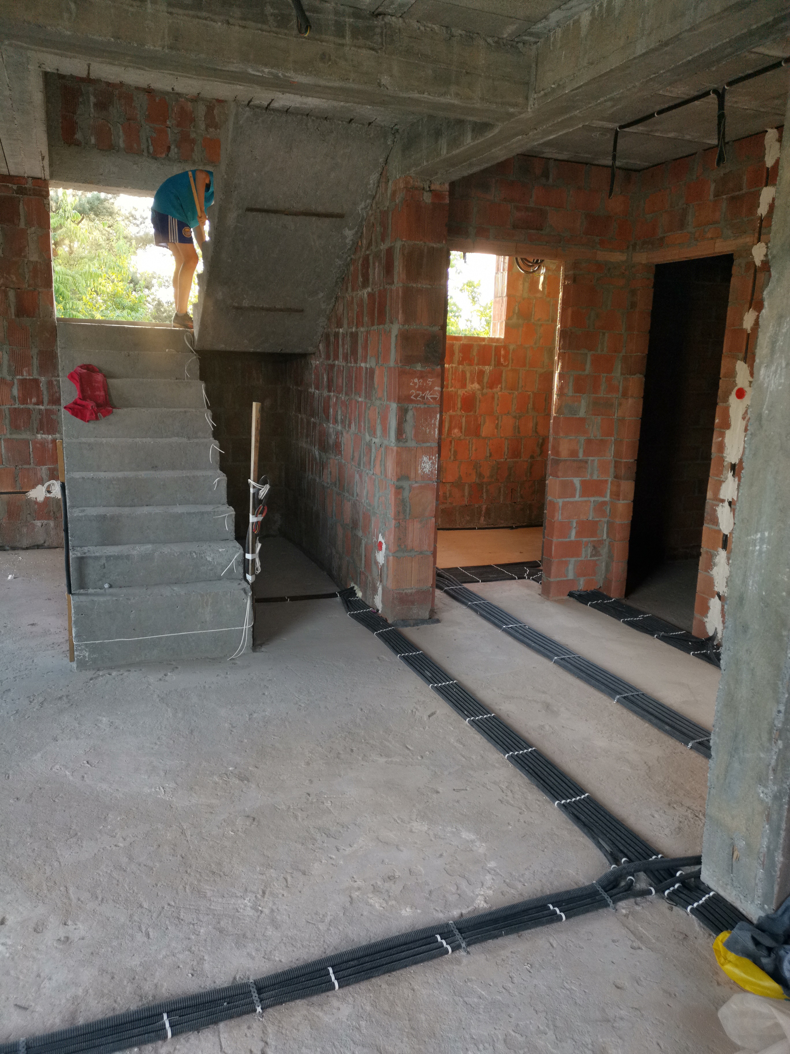 Wnętrze surowego budynku w trakcie prac instalacyjnych; widoczne kable w peszlach ułożone na podłodze, betonowe schody z czerwoną szmatką, osoba w oknie na schodach i surowe ściany z cegły.