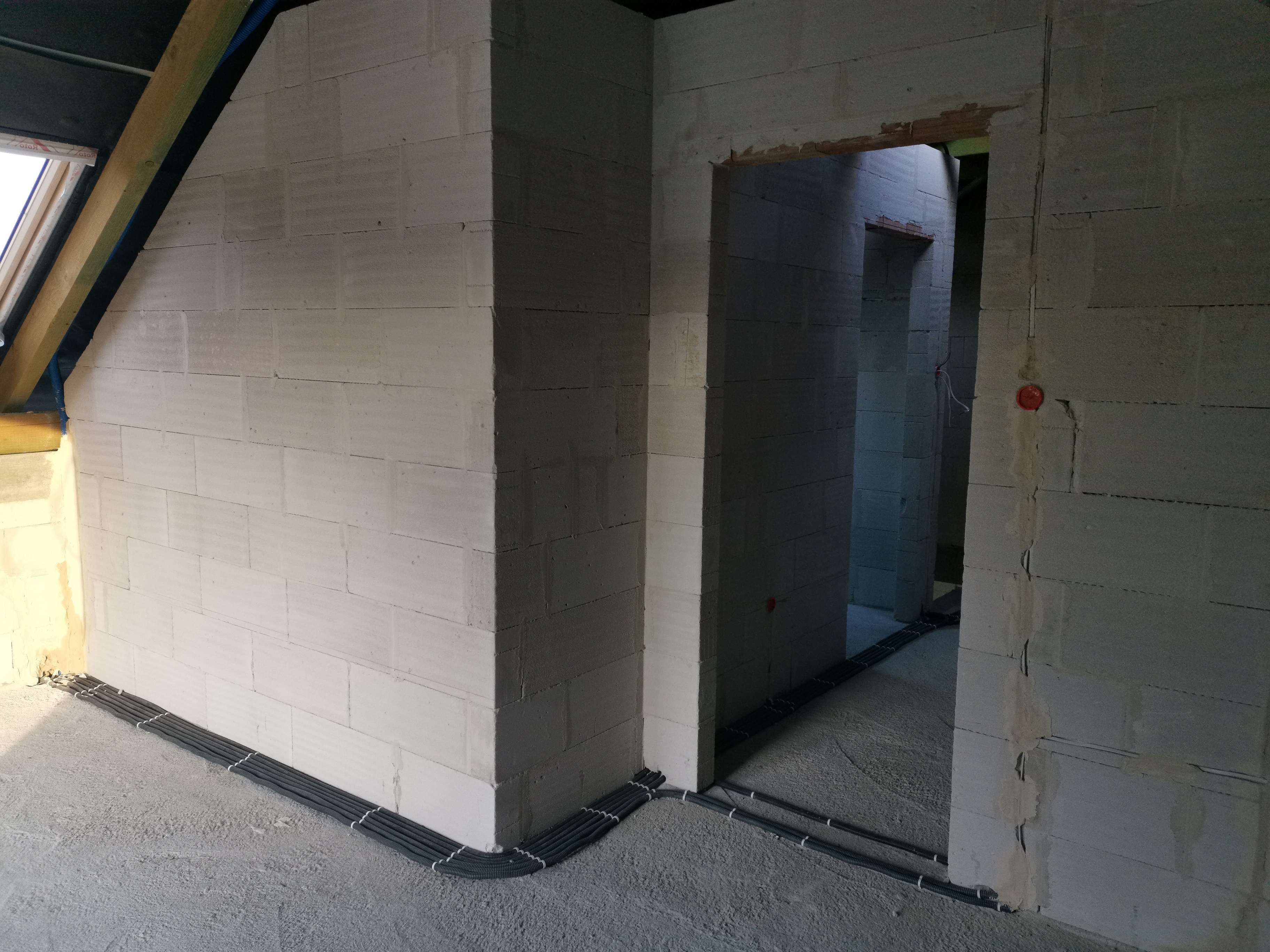 Surowy stan wnętrza w trakcie budowy, widoczne ściany z betonu komórkowego, kable elektryczne ułożone w peszlach przy podłodze, oraz otwór drzwiowy bez ościeżnicy.