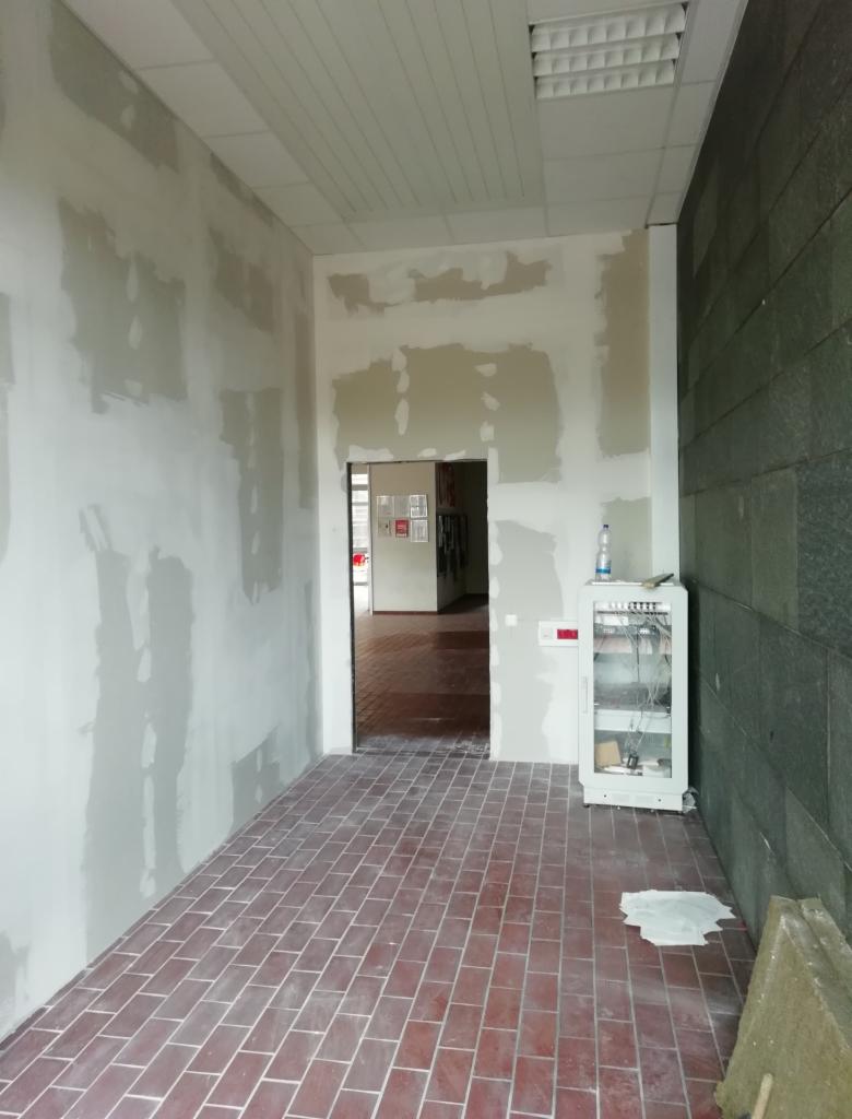 Wnętrze w trakcie prac wykończeniowych: ściany z widocznymi łączeniami płyt, przygotowane do dalszej obróbki, czerwona ceglana podłoga, biała szafka z przewodami i butelką wody, widok...