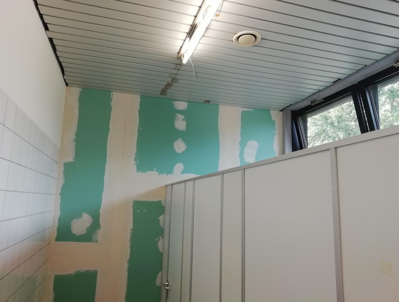 Wnętrze w trakcie remontu: częściowo położone płyty na ścianie, fragmenty zaszpachlowane, widoczny sufit z paneli i okno z widokiem na zieleń.