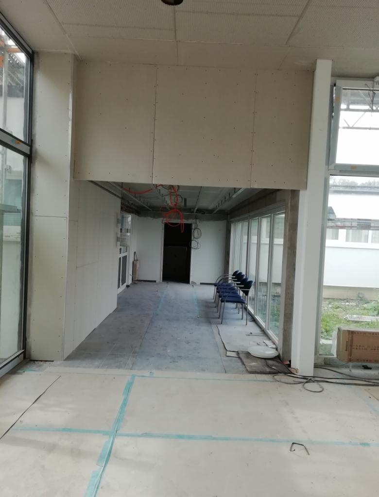 Wnętrze w trakcie prac wykończeniowych: montaż konstrukcji, widoczne okna, instalacje elektryczne pod sufitem, ślady taśmy malarskiej na podłodze.