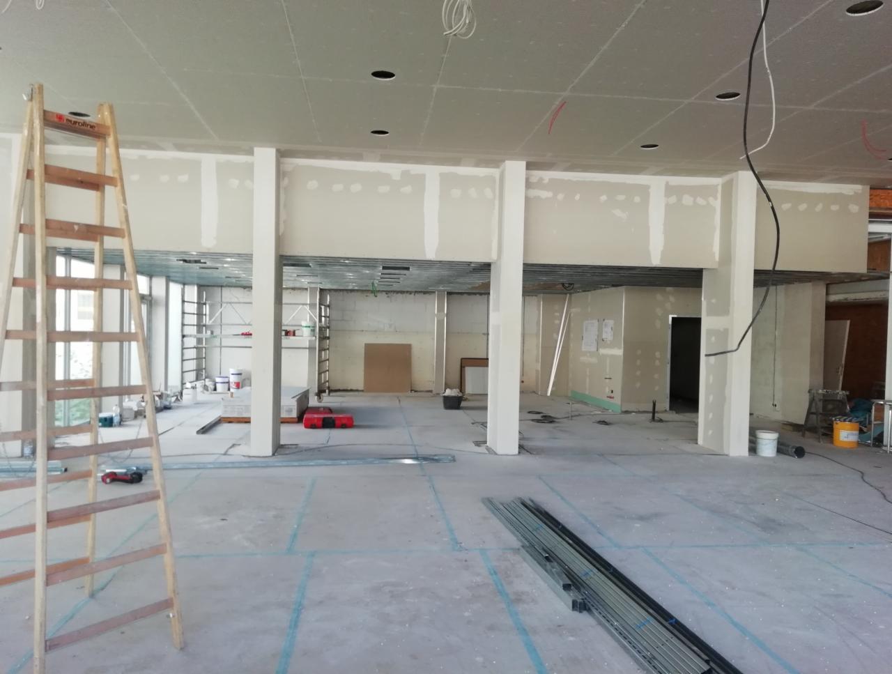 Wnętrze w trakcie prac wykończeniowych: widoczne konstrukcje sufitowe, częściowo zamontowane płyty, drabina malarska z lewej strony, narzędzia i materiały budowlane rozsiane po podłodze, surowe...