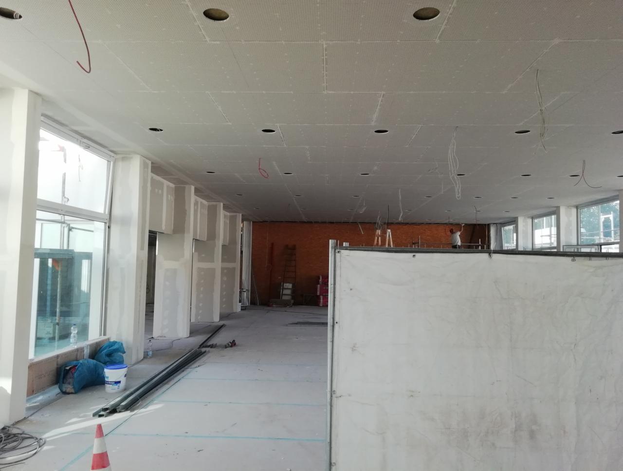 Wnętrze w trakcie remontu: widoczne profile, okna, częściowo zamontowane płyty na suficie z wyciętymi otworami na oświetlenie, instalacje elektryczne zwisające z sufitu.