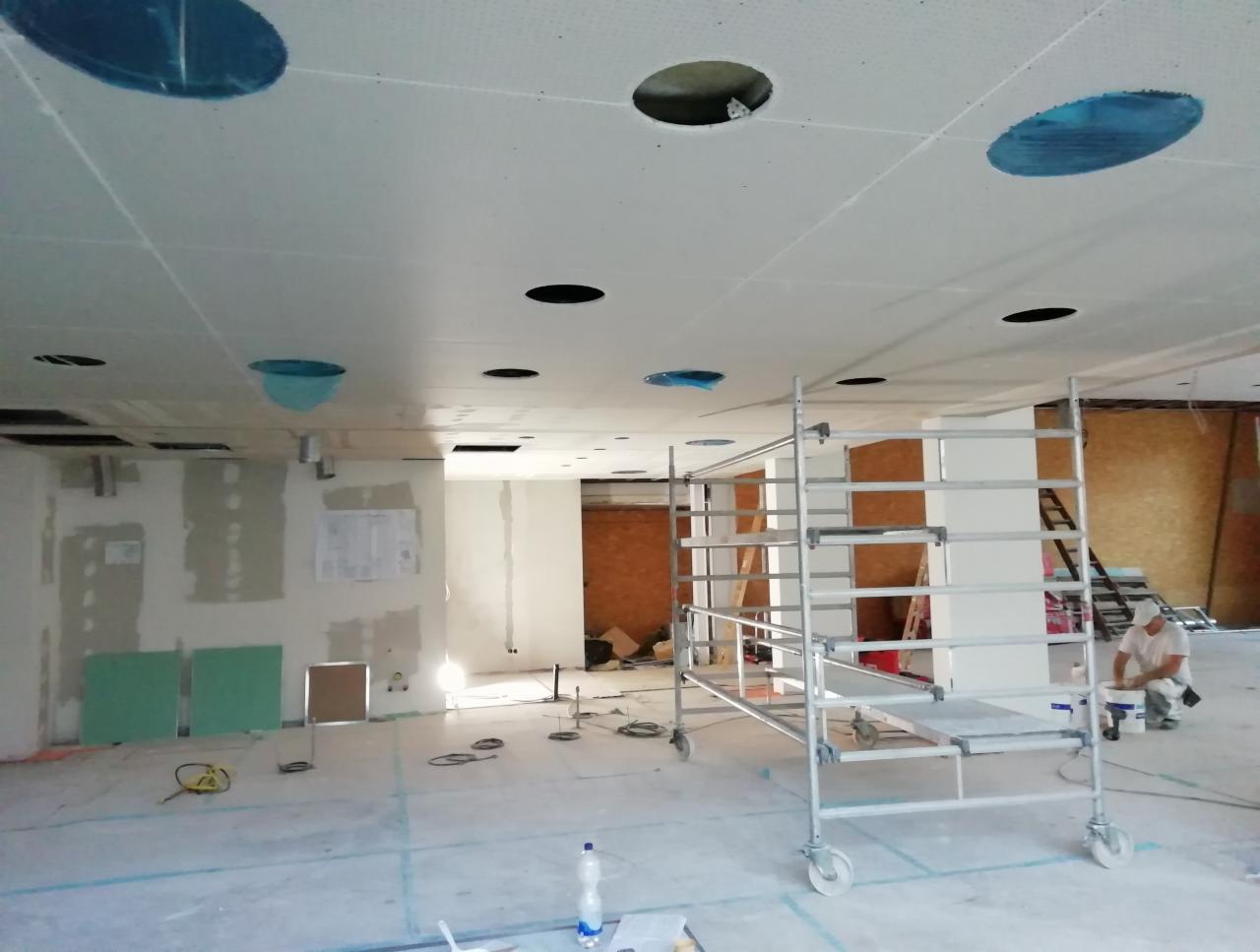 Montaż sufitu podwieszanego: widok na pomieszczenie w trakcie prac, z rusztowaniem, otworami w suficie zabezpieczonymi folią i pracownikiem przygotowującym farbę.