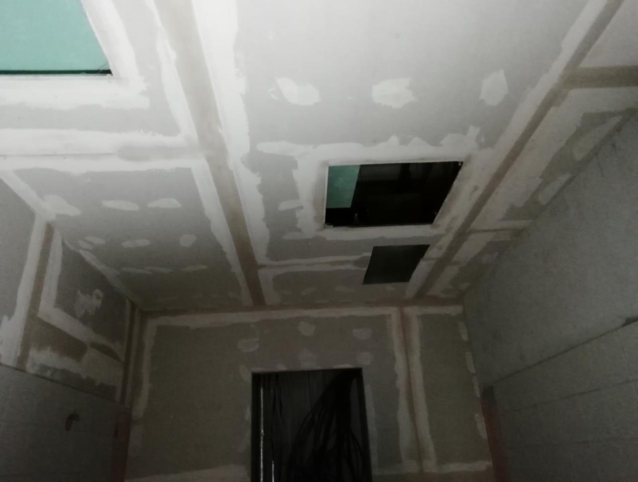 Wnętrze w trakcie prac wykończeniowych; widoczny sufit z otworami i ściany przygotowane do dalszych etapów, z widocznymi łączeniami i szpachlowaniem.