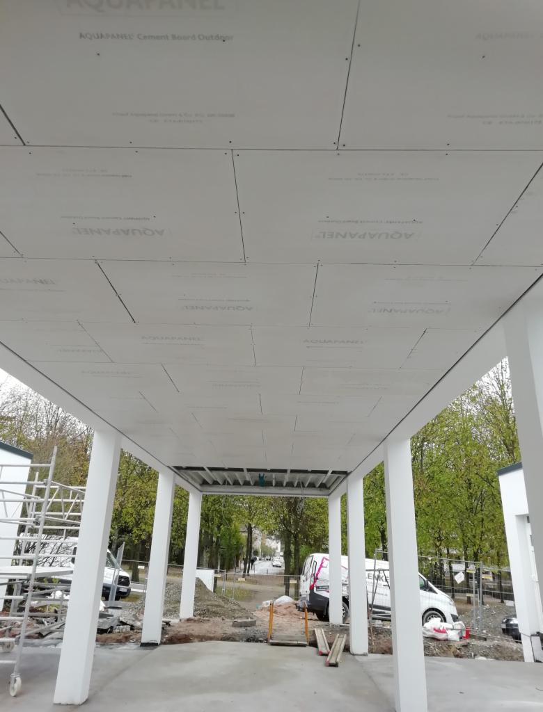 Montaż zewnętrznego zadaszenia z płyt cementowych Aquapanel, widoczne łączenia i wkręty, konstrukcja wsparta na białych kolumnach, w tle drzewa i zaparkowane samochody.