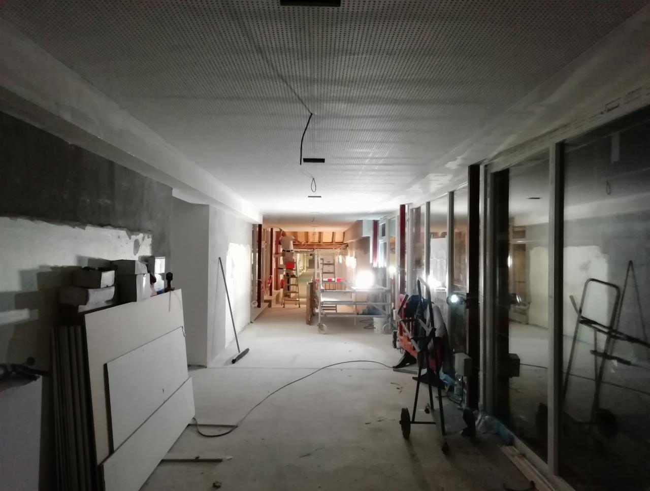 Długi korytarz w trakcie remontu, widoczne płyty na podłodze, częściowo otynkowane ściany, metalowe ramy okienne i perforowany sufit podwieszany z wystającymi przewodami oświetleniowymi.