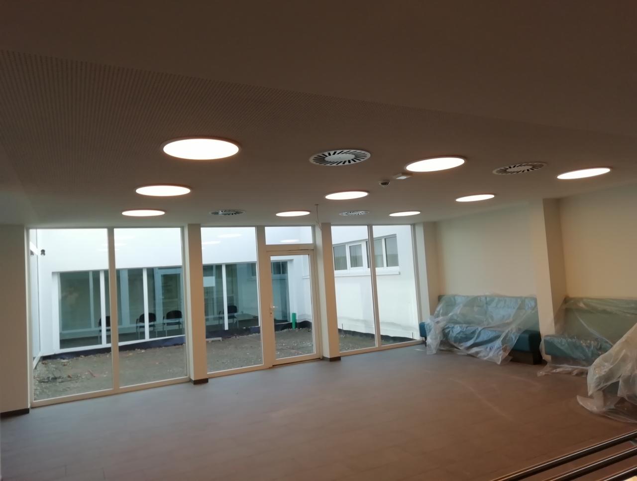 Wnętrze komercyjne z podwieszanym sufitem rastrowym, okrągłymi lampami i kratkami wentylacyjnymi, widoczne sofy zabezpieczone folią, duże okna z widokiem na dziedziniec.